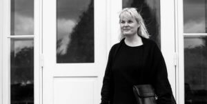 Black and white portrait of Rikke V. Stoltz – professional and calm expression, standing by a white door.