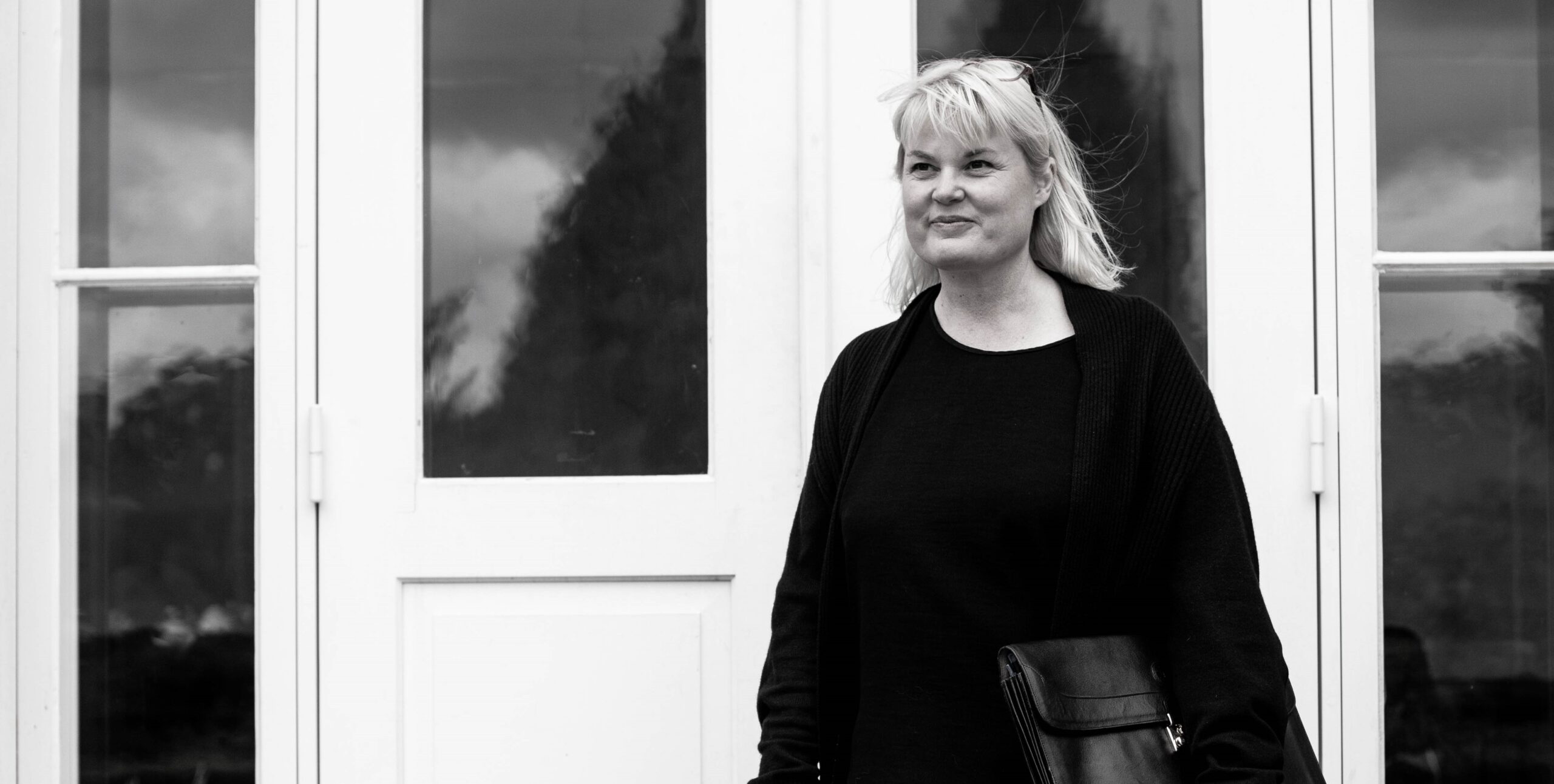 Black and white portrait of Rikke V. Stoltz – professional and calm expression, standing by a white door.